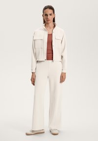 White cropped jacket with two chest pockets, paired with a fitted red top and wide-leg white pants. Features ribbed texture and neutral tones.