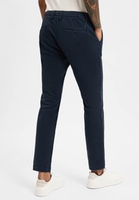 Navy blue tailored pants with a relaxed fit, elastic waistband, and two back pockets featuring button closures. Smooth cotton fabric, ankle length.