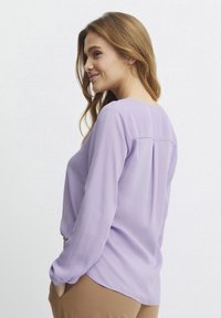 Helles Lavendel-Bluse mit langen Ärmeln, rundem Ausschnitt und Raffdetail auf der Rückseite. Weicher Stoff; lässige Passform. Kombiniert mit beigefarbenen Hosen.