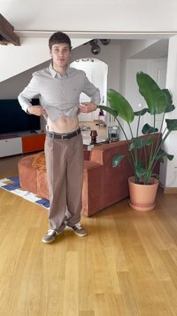 Jonge man die zijn gestreepte shirt optilt om zijn buik te tonen, staande in een woonkamer met een grote geplante pot en een oranje bank op een houten vloer.