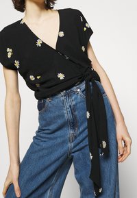 Top nero avvolgente con ricami di piccole margherite bianche e gialle, scollatura a v, maniche corte e vita legata, abbinato a jeans blu a vita alta.