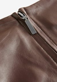 Fermeture éclair d'une veste en cuir marron, présentant une texture lisse et des éléments métalliques avec un tirette rectangulaire le long de la couture.