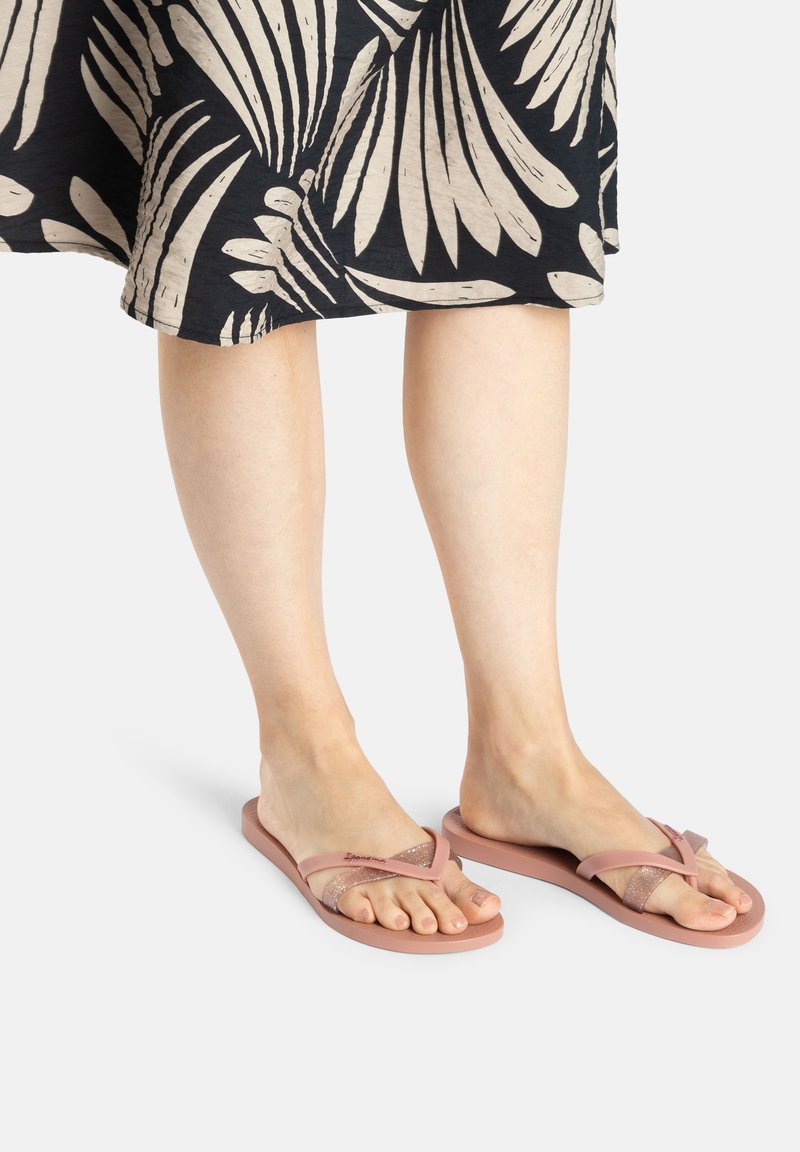 Pink flip-flops with a textured strap, featuring a glittery accent. Worn with a patterned black dress. Flat sole, open-toe design.