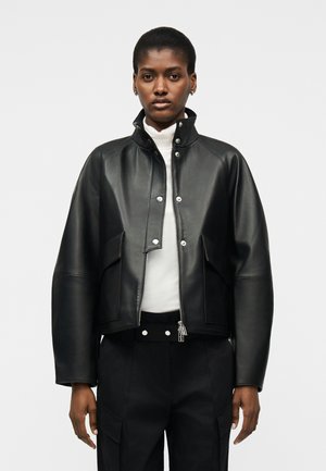 Γυναίκα που φοράει μαύρο δερμάτινο μπουφάν πάνω από ένα λευκό τοπ με ψηλό λαιμό και μαύρο παντελόνι, στέκεται μπροστά σε απλό ανοιχτό φόντο.