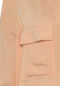 Light peach fabric with a smooth texture, featuring a patch pocket on the upper left sleeve and subtle woven detailing.