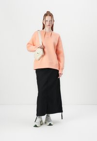 Sweatshirt laranja claro com decote redondo e ombros caídos, combinado com uma saia preta até ao tornozelo e ténis cinza-esverdeados. Pequena mala bege.