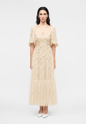 Jonge vrouw draagt een beige doorzichtige midi-jurk met bloemenborduursel en pofmouwen, staand met ontspannen armen langs het lichaam.