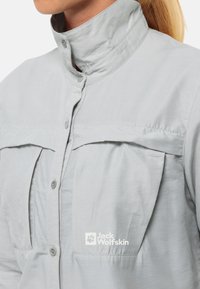 Hellgraues Hemd mit hohem Kragen, Knopfverschluss und zwei aufgesetzten Brusttaschen mit Knopf. Verfügt über das Jack Wolfskin-Logo auf der linken Seite. Glatte Textur.