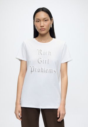 Kvinde med langt sort hår iført guldringe og en hvid t-shirt med teksten "Rich Girl Problems" i dekorativ skrift, stående mod en ensfarvet baggrund.
