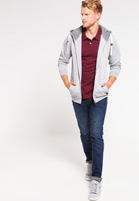 Veste à capuche gris zippée avec une capuche intérieure contrastante, portée sur un polo bordeaux, associée à un jean foncé et des baskets blanches.