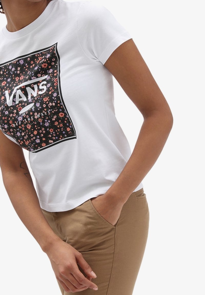 T-shirt blanc à manches courtes avec un graphique floral noir et le texte "VANS", en coton doux et de coupe ajustée. Un pantalon marron est visible.