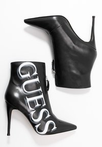 Botines de tacón alto de cuero negro con punta afilada y un logo "GUESS" plateado. Presentan una cremallera lateral y un acabado brillante y elegante.