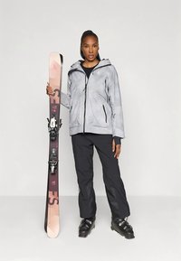 Woman in gray ski jacket, black pants, and ski boots holding a pair of pink and black skis upright against a gray background.