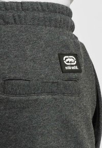Mørkegrå sweatpants med en struktureret stof, elastisk talje og en sort rektangulær logo-patch på baglommen.