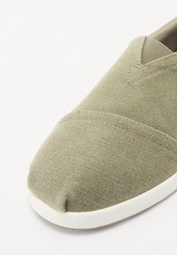 Zapatilla de tela verde tipo slip-on con un patrón de espiga texturizado, punta redondeada y suela de goma blanca. Diseño minimalista sin herrajes.