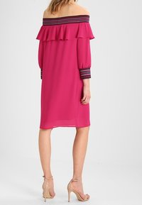 Un vestido rosa off-the-shoulder con un escote con volantes y mangas largas que presentan acentos a rayas en los puños. Tela suave y forma en A.