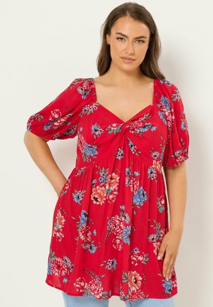 Kobieta ubrana w czerwoną sukienkę w kwiaty z bufkami na rękawach i dekoltem w serduszko, stojąca na jednolitym jasnym tle.