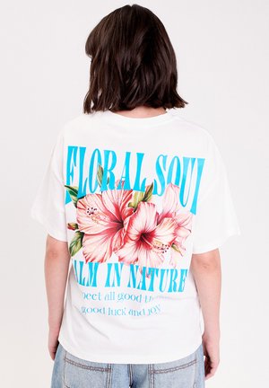 Personne aux cheveux foncés mi-longs portant un t-shirt blanc orné de grandes fleurs roses et d'un texte bleu indiquant « Floral Soul » et « Calm in Nature ».