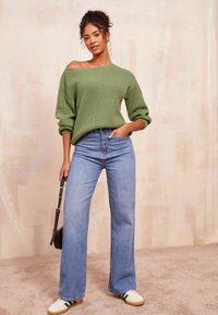 Grüner gerippter Pullover mit schulterfreiem Design, kombiniert mit hochtaillierten hellblauen weit geschnittenen Jeans und weißen Turnschuhen.