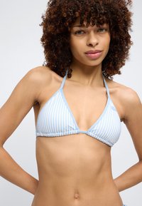 Γυναίκα με σγουρά μαλλιά που φοράει μπικίνι τοπ με ανοιχτό μπλε και λευκές ρίγες, στέκεται με τα χέρια πίσω από την πλάτη της μπροστά σε λευκό φόντο.