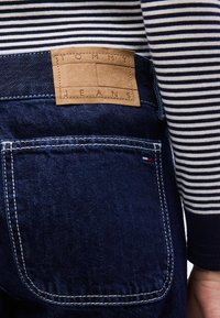 Πίσω όψη ατόμου που φορά σκούρο μπλε τζιν με δερμάτινη ετικέτα Tommy Jeans και μακρυμάνικο ριγέ πουκάμισο σε ασπρόμαυρο.