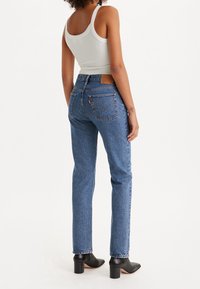 Jeans de mezclilla azul de tiro alto con un ajuste ligeramente holgado, que incluyen un parche de cuero de la marca, bolsillos traseros y un dobladillo sin terminar. Botas negras de tacón.