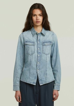 Donna con capelli lunghi e scuri, indossa una giacca di jeans azzurro chiaro abbottonata e pantaloni scuri, in piedi davanti a uno sfondo semplice.