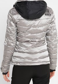 Vadderad jacka i glänsande silvermaterial med svart huva, horisontell sömnadsdesign och logodetalj på baksidan, lämplig för kallt väder.
