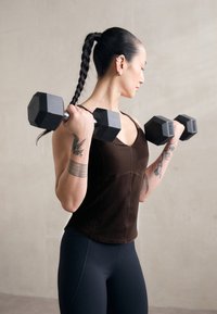 Crni heksagonalni utezi se drže u tamno smešnom uskom topu s V-izrezom, uparenim s tamnim tajicama. Vidljive su istaknute tetovaže na ruci.
