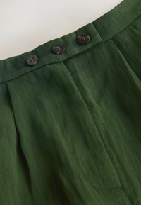 Groene stoffen rok van dichtbij, met een gladde textuur, plooien en drie donkere knopen bij de tailleband, wat een op maat gemaakte uitstraling benadrukt.