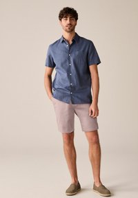 Rövid ujjú kék gombos ing, világos rózsaszín nadrág, és zöld slip-on cipő. Az ingnek van egy mellzsebe és laza szabása van.