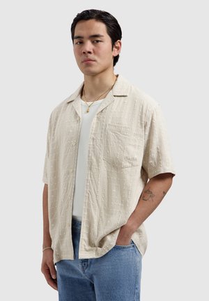 Young man with black hair, wearing a beige short-sleeve button-up shirt over a white tee and blue jeans, standing with one hand in pocket.