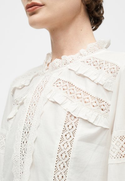 Κοντινή λήψη ενός ατόμου που φοράει λευκό μπλουζάκι με δαντελωτές λεπτομέρειες, βολάν στις άκρες και κεντημένα στοιχεία γύρω από τον λαιμό και τους ώμους.