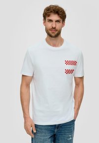 Camiseta de algodón blanca con escote redondo y mangas cortas, que presenta un patrón de cuadros rojos y blancos en el área del bolsillo del pecho izquierdo.