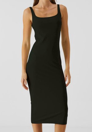 Femme portant une robe midi noire moulante sans manches avec un décolleté carré, debout devant un fond gris clair uni.