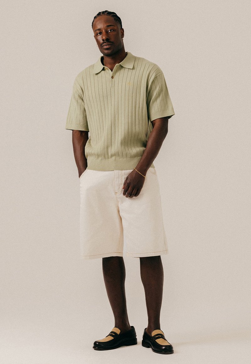 Homme aux cheveux tressés portant un polo côtelé vert clair, un short crème au genou et des mocassins noirs et beiges, debout les mains dans les poches.
