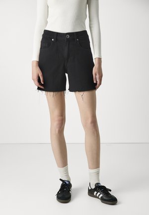 Pantalones cortos de mezclilla negra con cintura alta, dobladillo deshilachado y diseño de cinco bolsillos, combinados con una camiseta ajustada de manga larga blanca y zapatillas negras.