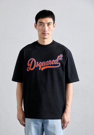 Man met kort zwart haar, gekleed in een zwart Dsquared2 t-shirt en lichtblauwe jeans, staand tegen een effen witte muur.
