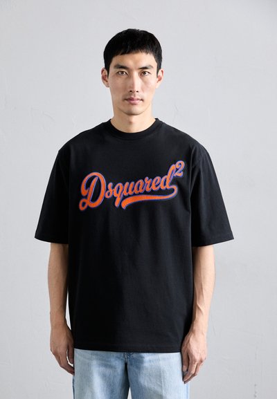 Uomo con capelli neri corti che indossa una t-shirt nera Dsquared2 e jeans azzurri chiari, in piedi contro un muro bianco semplice.