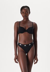 Vrouw staat rechtop met haar gezicht naar voren, gekleed in een zwarte bh en zwarte Adidas slip met logo tailleband tegen een neutrale achtergrond.