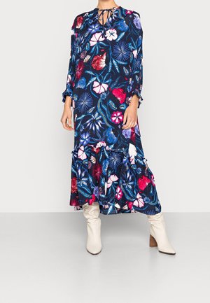 Vrouw draagt een lange marineblauwe bloemjurk met roze, rode en blauwe bloemen, gecombineerd met witte slouchy laarzen, staand tegen een eenvoudige achtergrond.