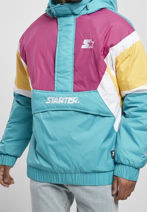 Homme portant une veste Starter colorée avec des panneaux turquoise, violet, jaune et blanc, des poignets élastiques, et un logo sur la poitrine et la poche avant.