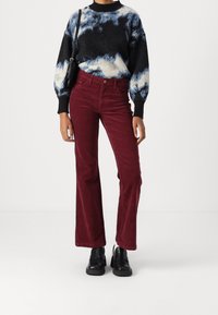 Rote Corduroy Schlaghosen kombiniert mit einem schwarz-blauen Batik-Pullover. Eine schwarze Umhängetasche und schwarze Schuhe runden das Outfit ab.