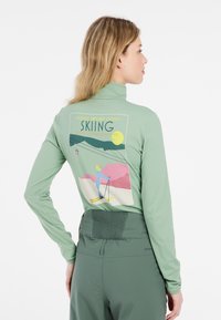 Zielona bluzka z długim rękawem, z grafiką narciarską na plecach, o wysokim kołnierzu i teksturowanej tkaninie, w komplecie z dopasowanymi zielonymi spodniami.