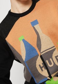 Tröja med svarta och orange sektioner som har en grafik av två dryckesflaskor, en med en blå etikett och den andra med en grön etikett.