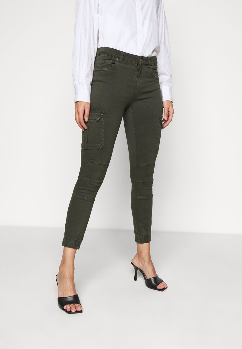 Pantalon cargo vert olive avec poignets élastiques aux chevilles, deux poches latérales et une ceinture ornée. Associé à des sandales à talons noires.