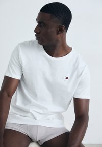 Hombre sentado vistiendo una camiseta blanca de manga corta y calzoncillos tipo bóxer gris claro, mirando hacia su izquierda sobre un fondo blanco liso.