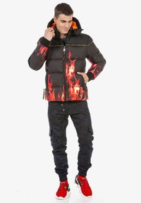 Schwarze Pufferjacke mit rotem Flammenmuster am Saum, ausgestattet mit einem orangefarbenen Kapuzenfutter, einem Reißverschluss und elastischen Bündchen. Kombiniert mit dunklen Camouflage-Hosen.
