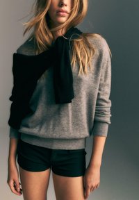 Giovane che indossa un maglione grigio a maniche lunghe con un maglione nero drappeggiato sulle spalle e pantaloni corti neri su uno sfondo neutro.