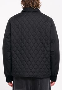 Chaqueta negra acolchada con un patrón de diamantes en la parte posterior, cuello alto y puños acanalados. La tela es suave y tiene un ligero brillo.